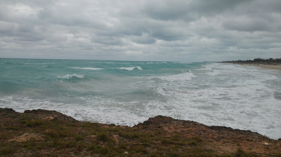 Grey sky over rough turquoise ocean waves breaking onto a rocky shoreline, with a long sandy beach stretching into the distance.