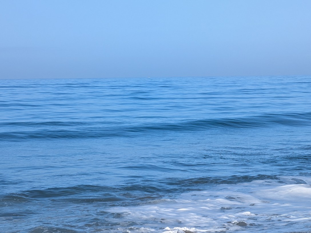 Calm ocean surface with soft blue tones and a gentle wave, horizon disappearing into haze