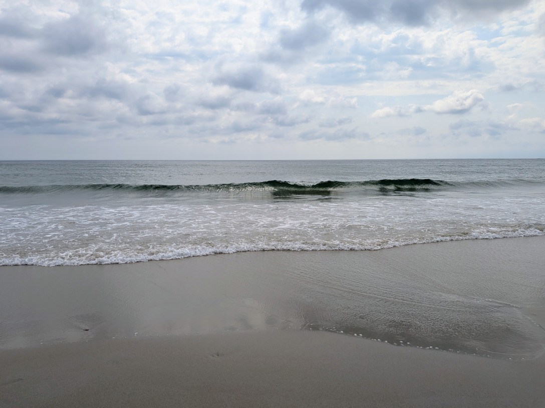 A single wave breaking against a calm grey shoreline under an overcast sky, the horizon line level behind it.